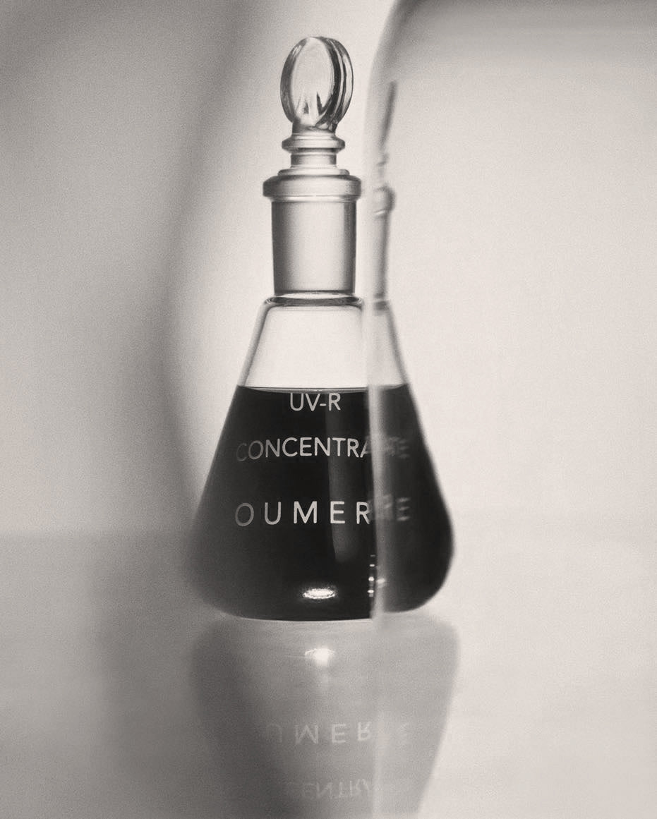 Black and white image of a glass bottle labeled 'OU-MERE' with a dropper on a light background
