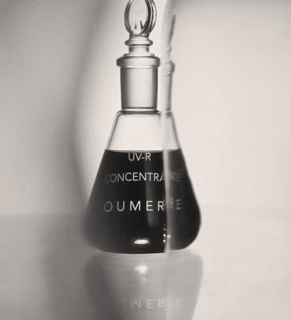 Black and white image of a glass bottle labeled 'OU-MERE' with a dropper on a light background